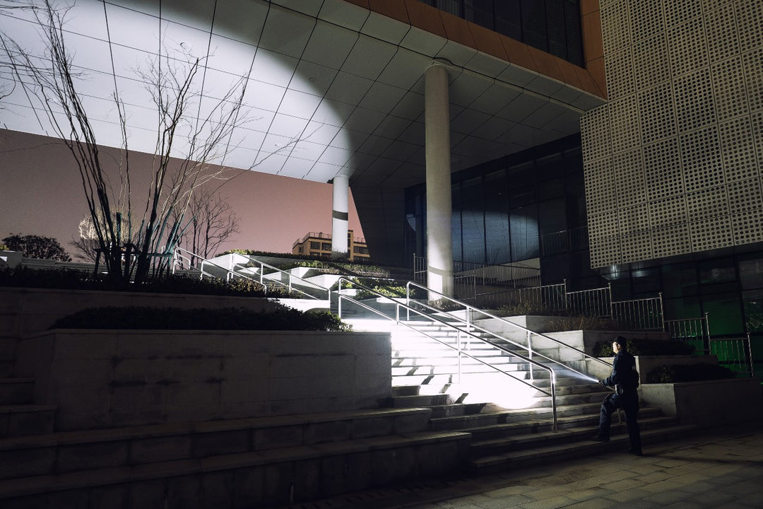 A security guard using the Fenix TK15R adaptive flashlight on a staircase in front of a modern building 